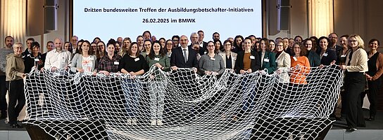Eine große Gruppe Menschen stehen vor einer Beamerleinwand und halten ein großes Netz in den Händen